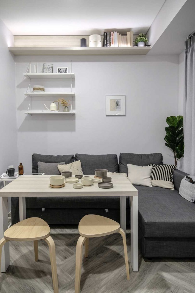 The little living room can be easily turned into a dining space, there's a grey sectional, a table and stools and a shelf with lights