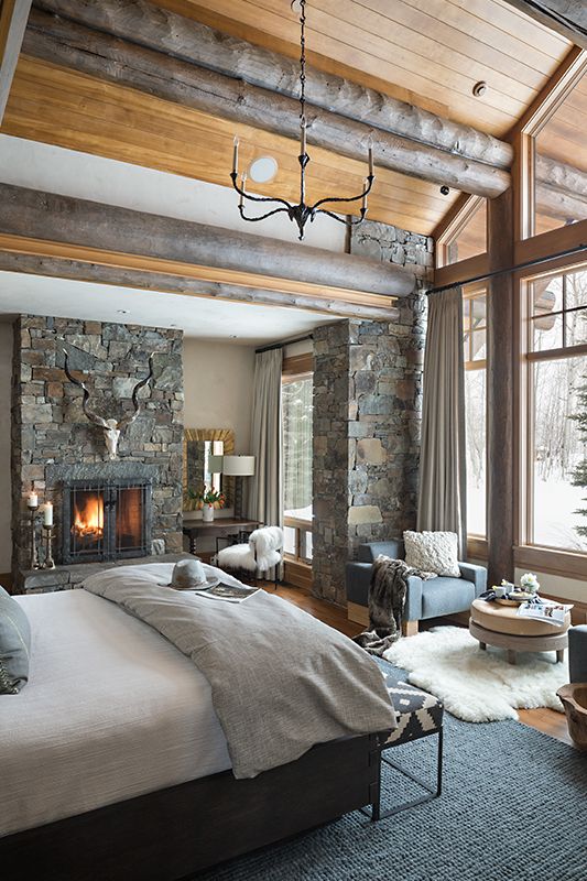 a rustic bedroom with wooden logs on the ceiling, a pillar clad with stone and a stone fireplace