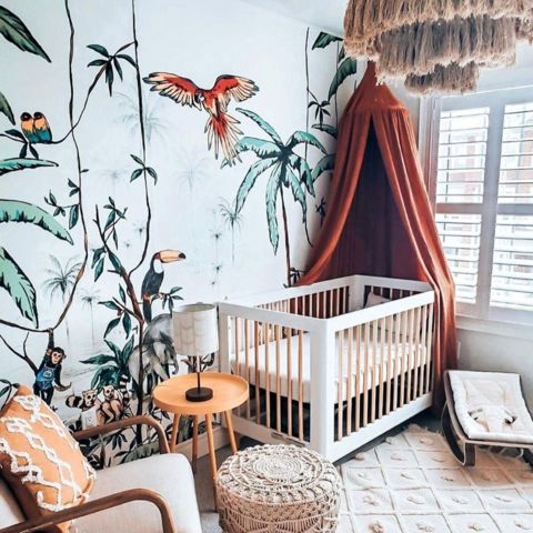 a jungle themed nursery with tropical wallpaper, a rust colored canopy and a boho tassel chandelier plus a boho ottoman