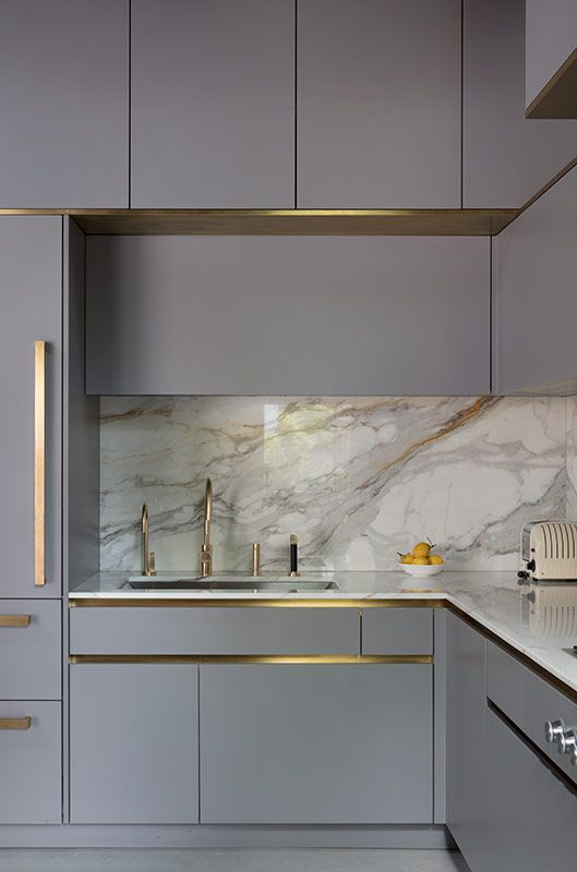 a beautiful grey kitchen with built-in lights, a white marble backsplash and countertops plus gold fixtures