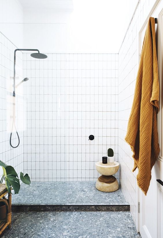 a chic and simple bathroom with white tiles, a grey terrazzo floor, mustard textiles and potted plants plus black fixtures