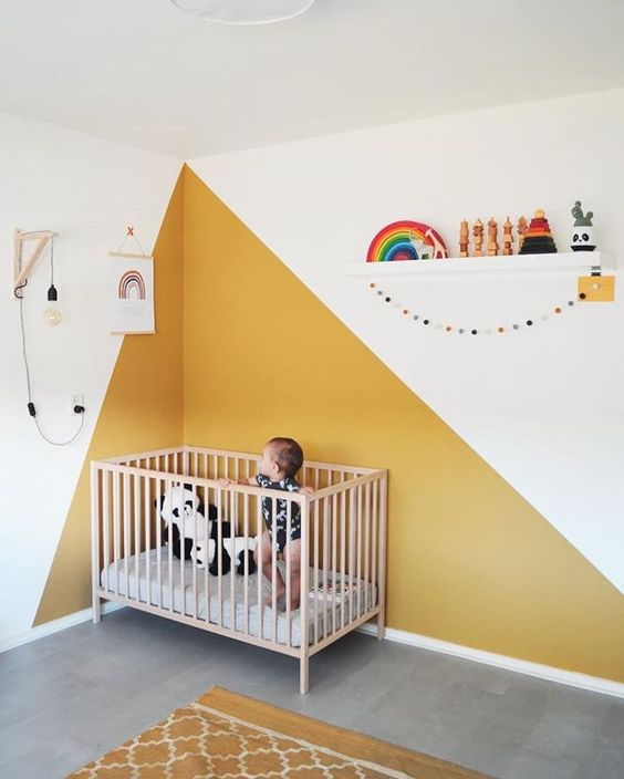 a bright modern nursery with yellow color blocking on the wall and a yellow rug, a neutral crib, a shelf with toys and some posters
