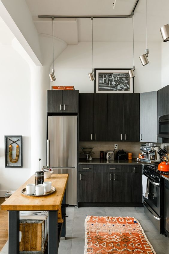 a black L-shaped kitchen with a tile backsplash, a small kitchen island with a butcherblock countertop and pendant lamps