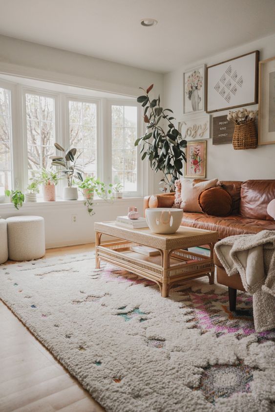 a boho living room with a leather sofa, a rattan table, a gallery wall and potted plants filled with light through a bow window