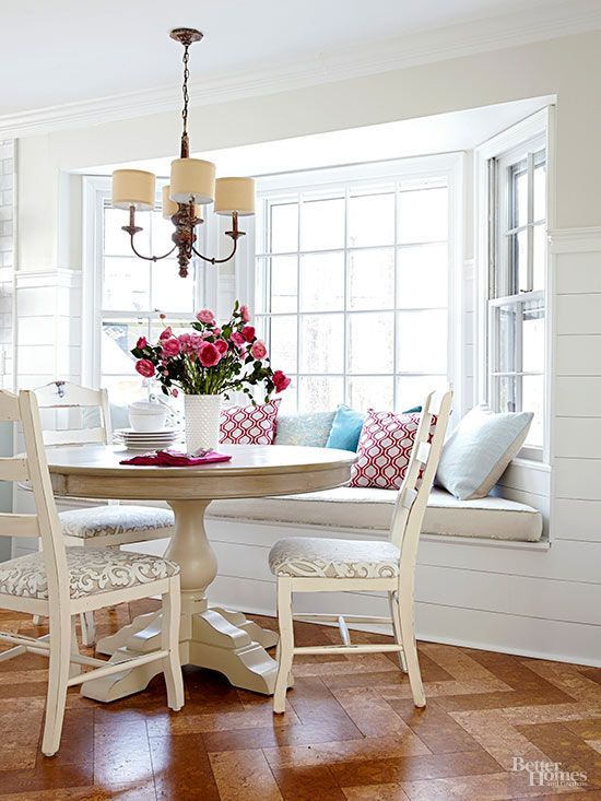 a charming cottage dining space with a bow window, a windowsill seating, a round table and neutral chairs plus a vintage chandelier