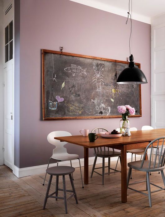 a chic dining room with a mauve accent wall, a wooden table and mismatching chairs, a chalkboard and a pendant lamp