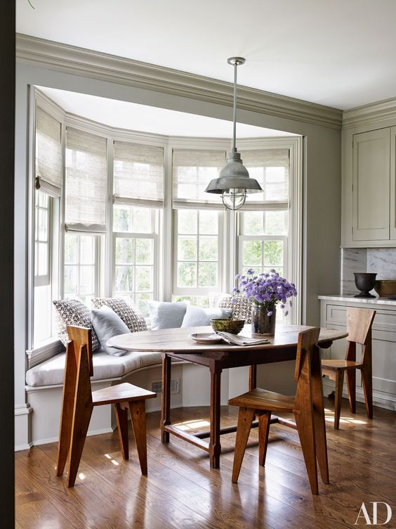 a cozy farmhouse nook with a bow window, a windowsill banquette seating, a wooden table and wooden chairs plus a retro lamp