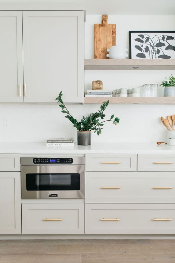 a dove grey kitchen with shaker cabinets, a white backsplash and countertops, floating shelves and gold handles is very elegant