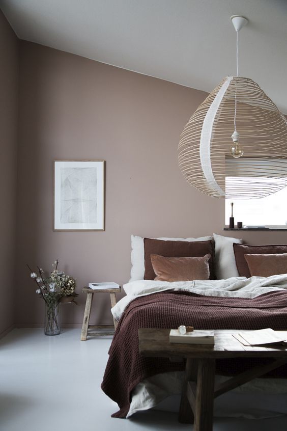 a mauve bedroom with simple furniture, layered bedding, an artwork, a rattan lamp and some blooms is very soothing