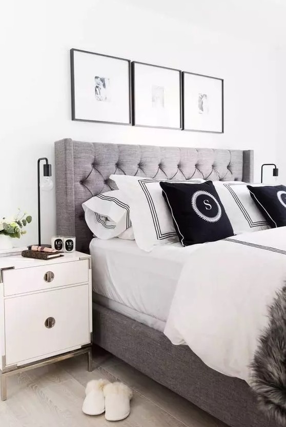 a modern glam bedroom with a grey upholstered bed with black and white bedding, a neutral nightstand with brass legs and a grid gallery wall