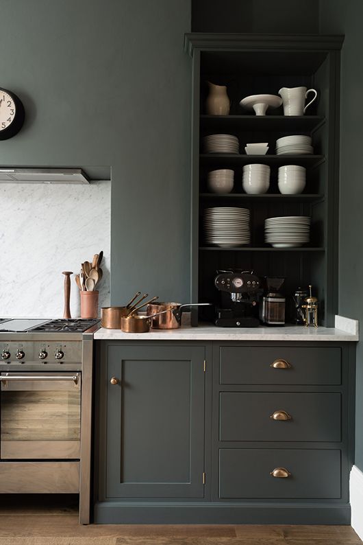 a monochromatic dark grey kitchen with shaker style cabinets, open compartments and drawers, a white backsplash and countertops and brass touches