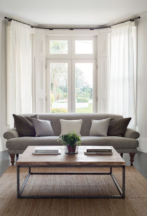 a neutral farmhouse living room with a bow window draped with curtains for more privacy and less light and a grey sofa