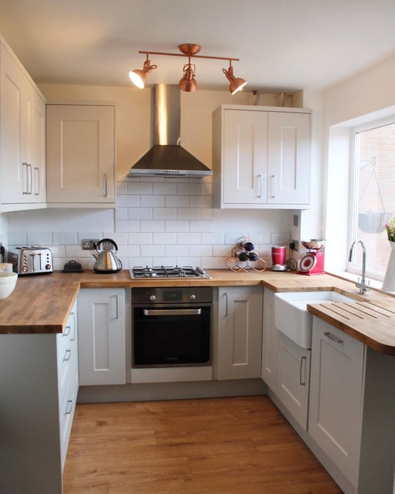 a small and comfy U-shaped kitchen with white cabinets, butcherblock countertops, a white tile backsplash and copper lamps