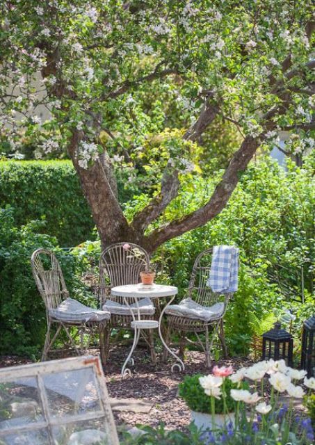 a small and cozy dining space under a blooming tree, with vintage rattan chairs and a mini table, with candle lanterns and potted blooms
