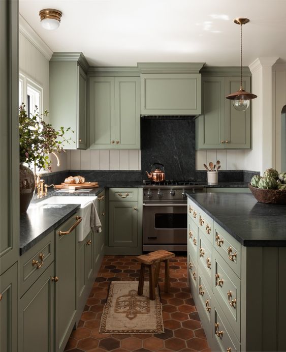 a vintage sage green kitchen with shaker style cabinets, black quartz countertops and a backsplash, gilded and brass fixtures