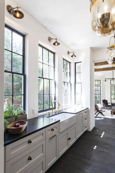 a vintage white one wall kitchen with black countertops, large windows, brass sconces and elegant pendant lamps