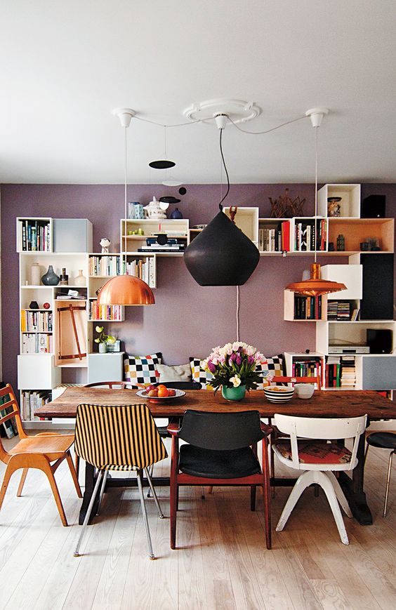 an eclectic dining room with a mauve accent wall, lots of open shelves, a banquette seating, a wooden table and mismatching chairs