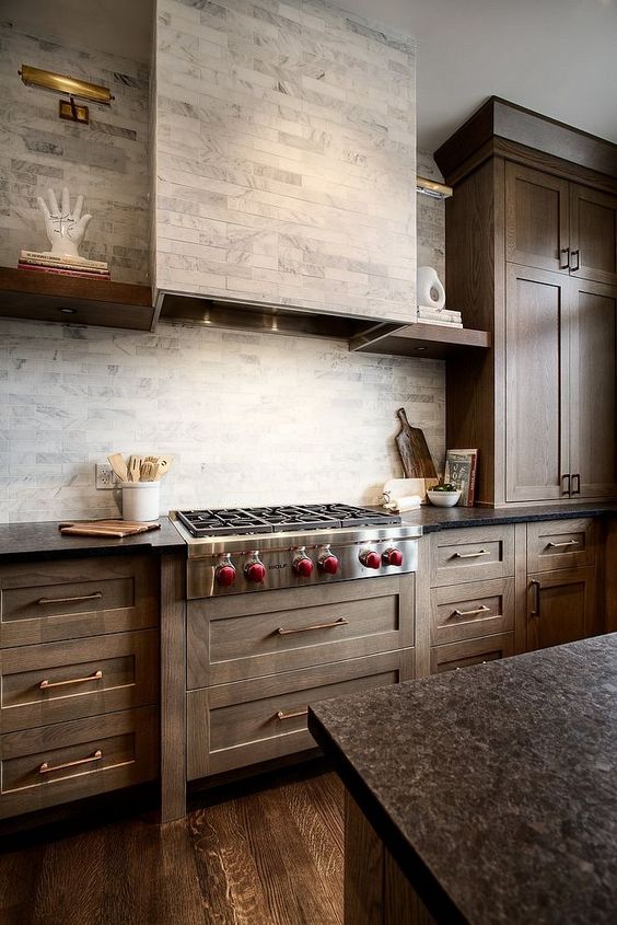 a farmhouse kitchen with stained cabinets, a grey marble backsplash and a hodo clad with the same tiles for a seamless look
