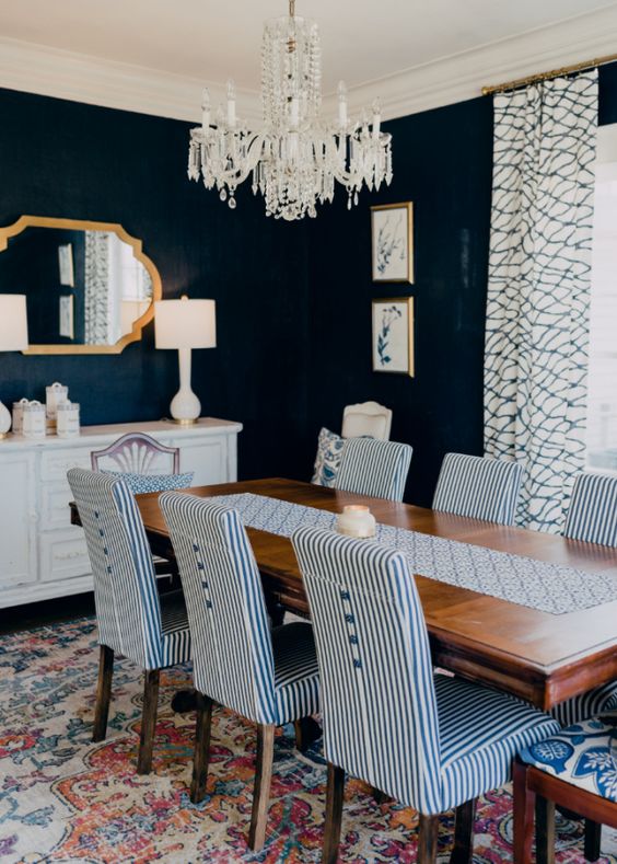 a bold dining space with navy walls, a white sideboard, a wooden table, striped chairs, a white crystal chandelier and a printed rug