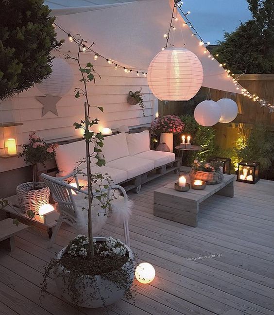 une terrasse accueillante avec des meubles modernes en palettes et en rotin, des lanternes à bougies, des guirlandes lumineuses et des lampes en papier dans tout l'espace