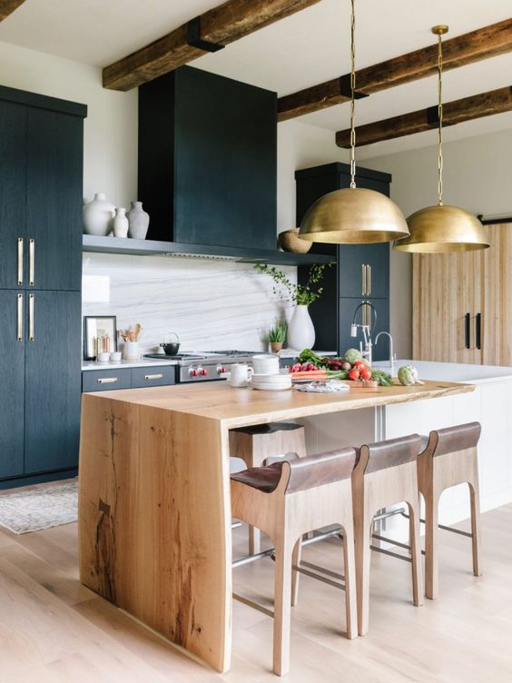 a beautiful kitchen with black cabinets, a white stone backsplash, a live edge blonde wood countertop and matching stools plus gold lamps