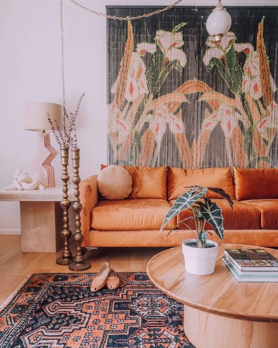 a boho living room with a floral tapestry, an orange sofa, a round table and a side table plus candle holders and a boho rug