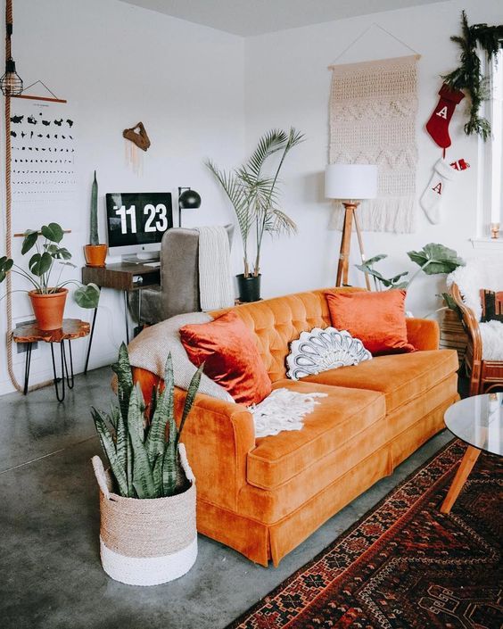 a boho living room with an orange sofa, potted plants, a macrame hanging, a small workspace in the corner is amazing