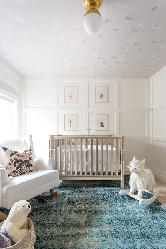 a chic neutral nursery with paneled walls, a grey crib, a white rocker, a basket with toys and a lovely printed ceiling