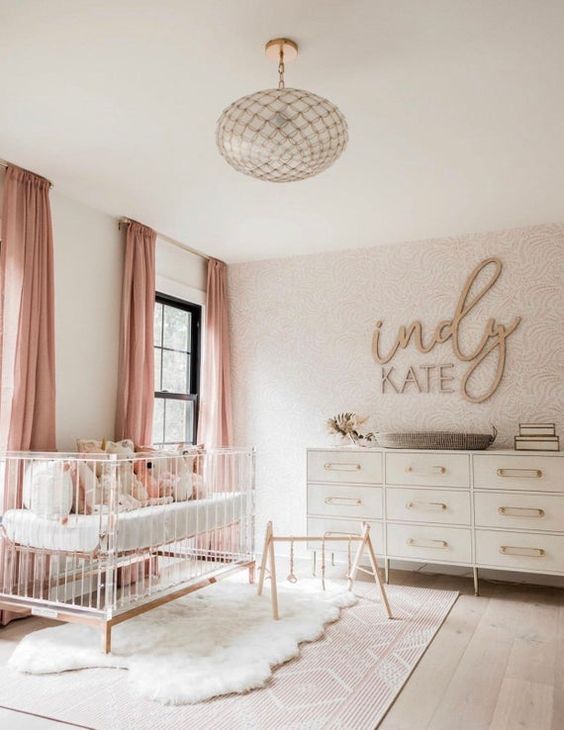 a lovely modern nursery with a wallpaper wall, a neutral dresser and an acrylic crib, a beautiful glass pendant lamp and pink curtains