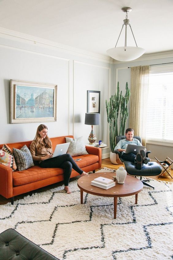 a mid-century boho living room done in neutrals, with an orange sofa and a black leather chair plus a statement cactus and artworks