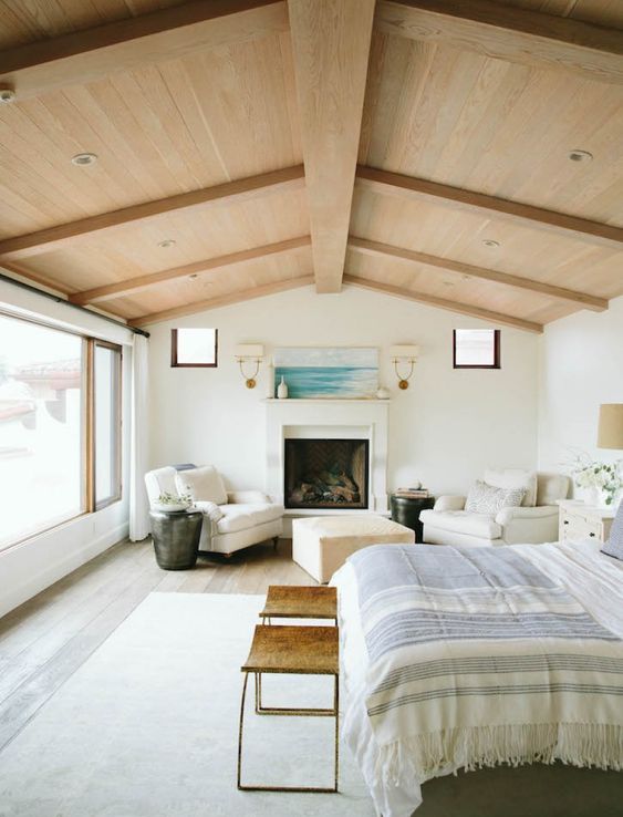 a neutral and beautiful bedroom with a panoramic window, a blonde wood ceiling with beams, a fireplace, white chairs and an ottoman, a bed and leather stools