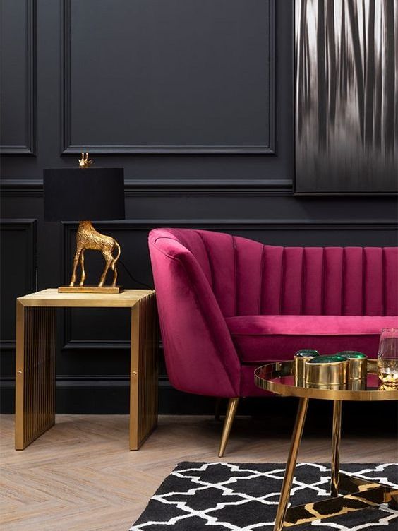 a refined moody space with black paneled walls, a fuchsia sofa, a gold side and coffee table, a creative table lamp and a printed rug