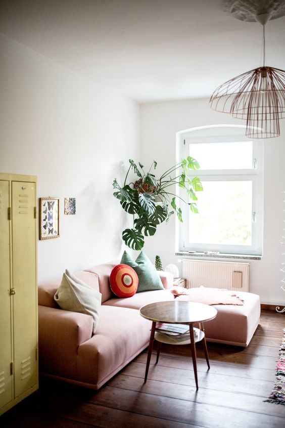 a small bright living room with a yellow locker, a low pink sofa, some potted plants and a catchy pendant lamp