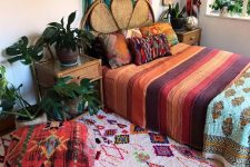a boho maximalist bedroom with a rattan bed and wooden nightstands, a bold rug and bedding, a bunting and hats for decor on the wall