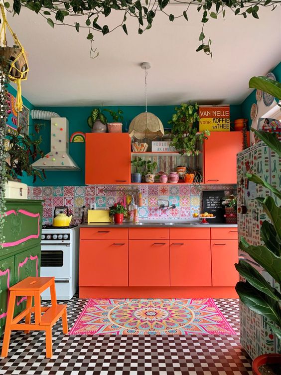 a bright maximalist kitchen with coral furniture, a bright floor and rug, a green wall and a colorful backsplash plus lots of potted plants