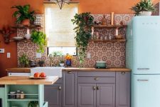 a bright maximalist kitchen with grey cabinets, an aqua-colored kitchen island and a matching fridge, rust walls and bright tiles