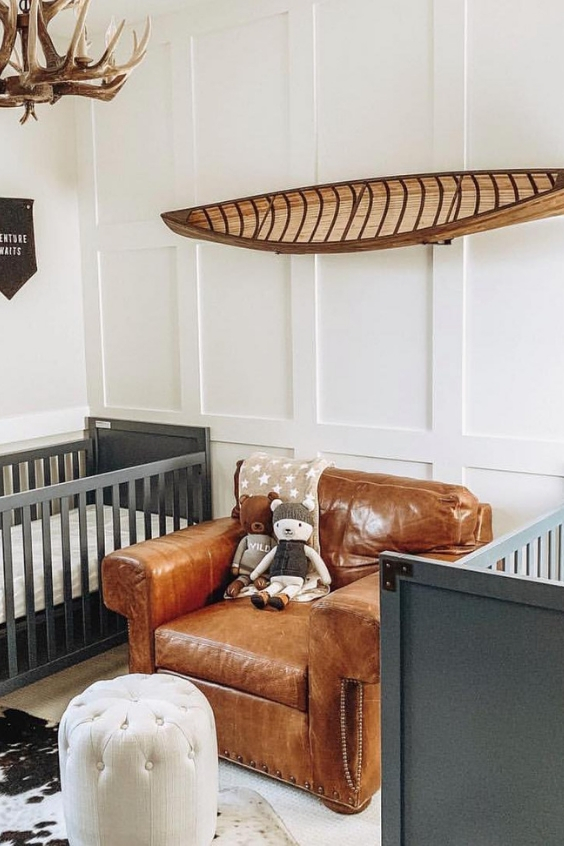 a catchy adventurous twin nursery with grey cribs, a leather chair, a canoe, an antler chandelier and white paneled walls