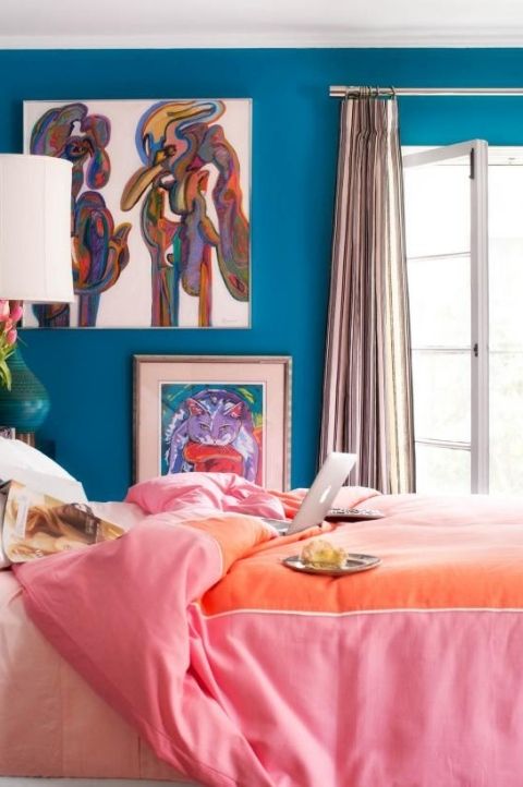 a colorful maximalist bedroom with blue walls, bright artworks, colorful bedding and curtains plus a green table lamp