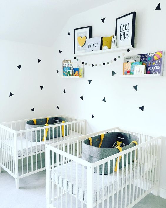 a contemporary twin nursery with white cribs, shelves with art and books and catchy decals for an accent