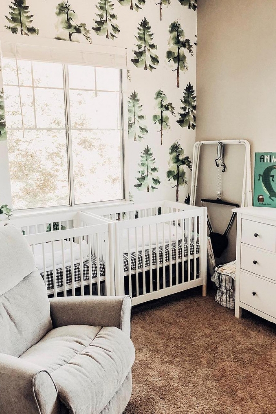 a forest-themed twin nursery with white cribs, tree-printed wallpaper, white and grey furniture and a pretty soft rug