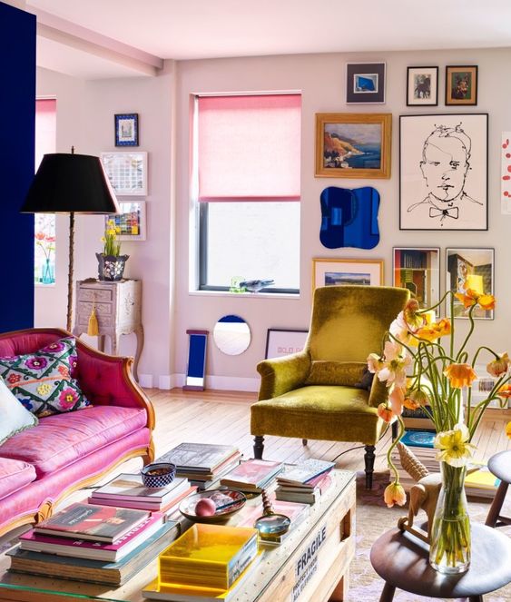 a lovely maximalist living room with blush walls, a hot pink sofa, a mustard chair, a coffee table with lots of books and a bright gallery wall