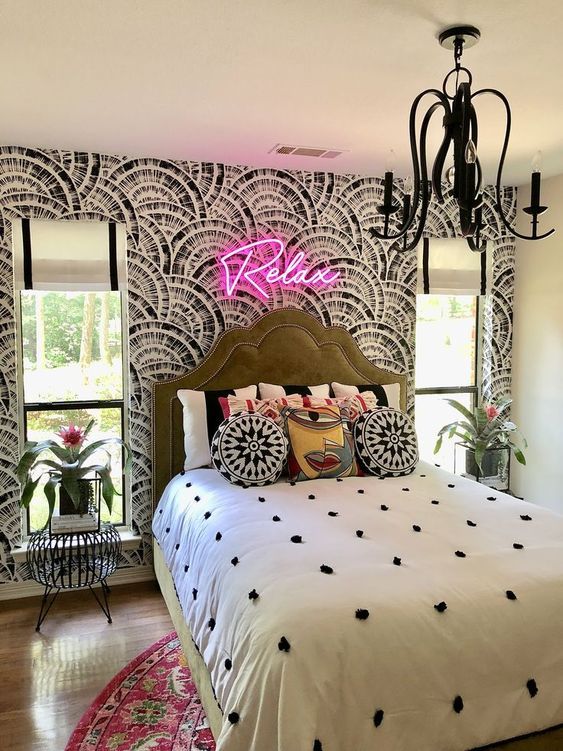 a maximalist bedroom with a printed accent wall, a green bed, bright bedding and a rug, a neon light and a black chandelier