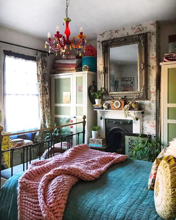 a maximalist bedroom with floral walls, a metal bed, bright textiles, a colorful chandelier, a fireplace and a yellow dresser