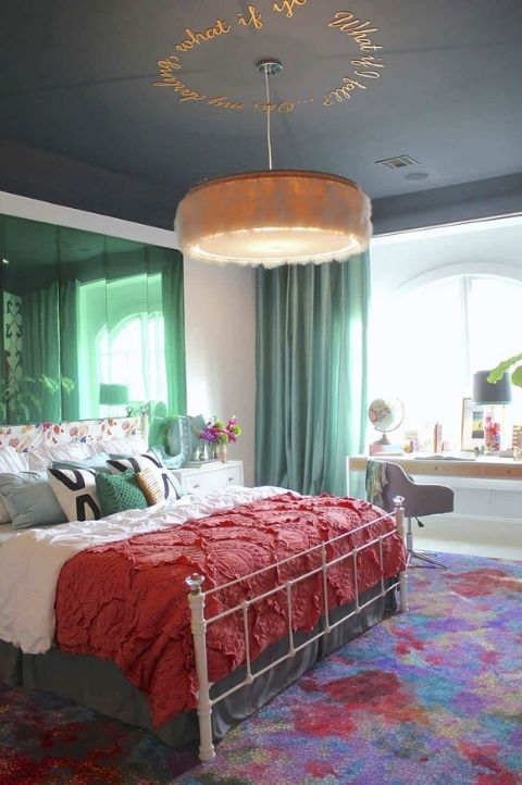 a maximalist bedroom with simple furniture, green mirrors and curtains, a colorful rug and bold bedding plus a fluffy chandelier