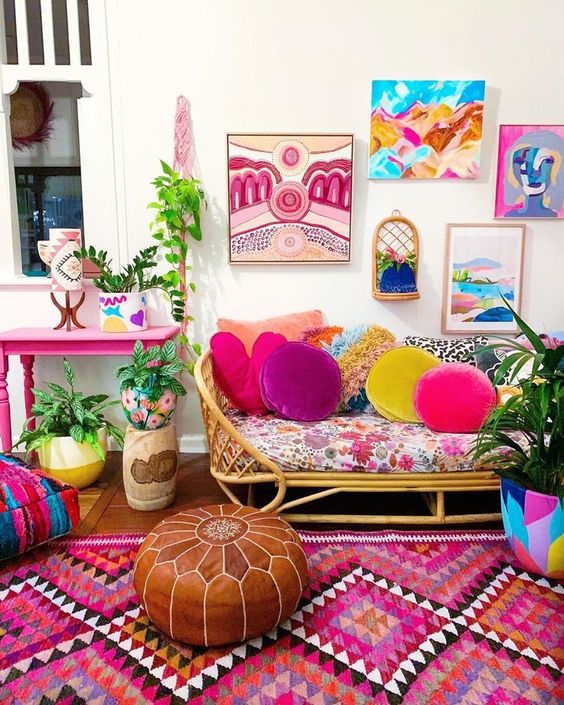 a maximalist space with a hot pink table, a rattan sofa, colorful textiles and pillows, ottomans and a gallery wall is wow