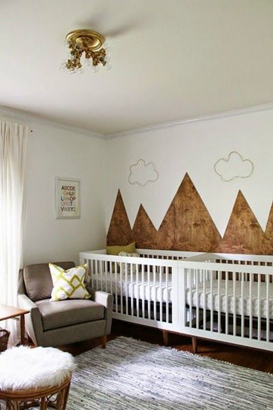 a mountain twin nursery with an accent mountain wall, white cribs, a grey chair, a stained table and a stool is welcoming