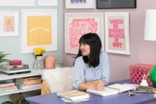 a pastel maximalist home office with a colorful gallery wall, a lilac desk, a hot pink stool and pastel books and a lamp