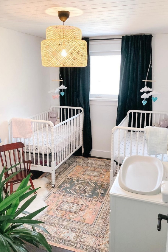 a small and cute twin nursery with white cribs, navy black-out curtains, a printed rug, potted plants and a lovely retro lamp