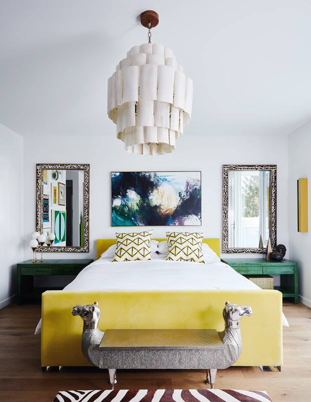 a stylish maximalist bedroom done in white, with a yellow upholstered bed, green nightstands, mirrors, a statement artwork and a catchy chandelier