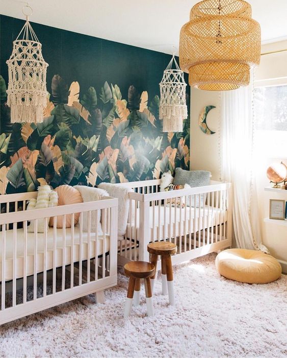 an eye-catchy boho twin nursery with a tropical leaf accent wall, white cribs, wooden stools, macrame and a pendant lamp is wow